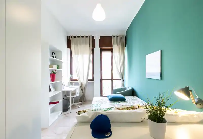 Bright, modern bedroom with a double bed against a teal accent wall, a desk by the window and cozy textiles and plants.