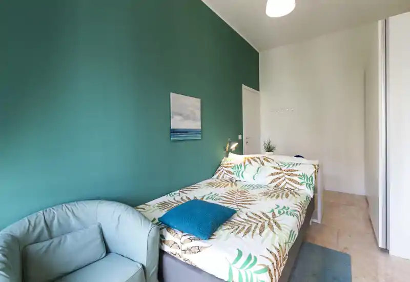 Camera da letto luminosa e di stile con letto matrimoniale, biancheria a motivo tropicale, parete accent color verde petrolio e una comoda poltrona — perfetta per soggiorni rilassanti.