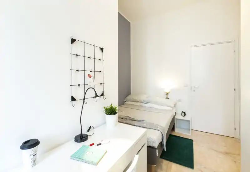Bright, minimal bedroom with a neatly made double bed, bedside lamp and a small desk in the foreground — ideal for single or couple stays.