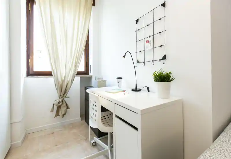 Small, bright bedroom corner with a white desk by a sunlit window, practical chair, desk lamp and a potted plant — ideal for work or study.