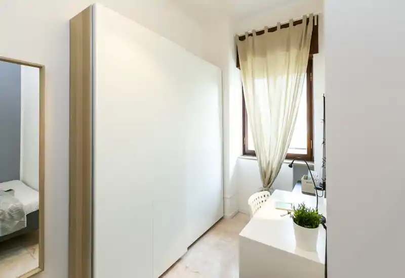 Bright, minimal bedroom corner with a white wardrobe, small desk by the window and a glimpse of the bed seen through the open doorway; clean modern styling and natural light.