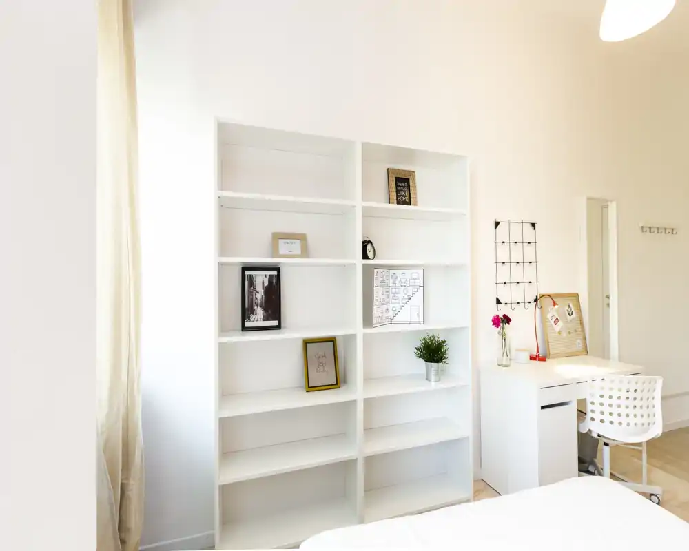 Angolo camera luminoso e minimal con una libreria bianca e una piccola scrivania. Si vede una porzione del letto in primo piano, scaffali con cornici e una pianta, e un'area di lavoro ariosa e ordinata.