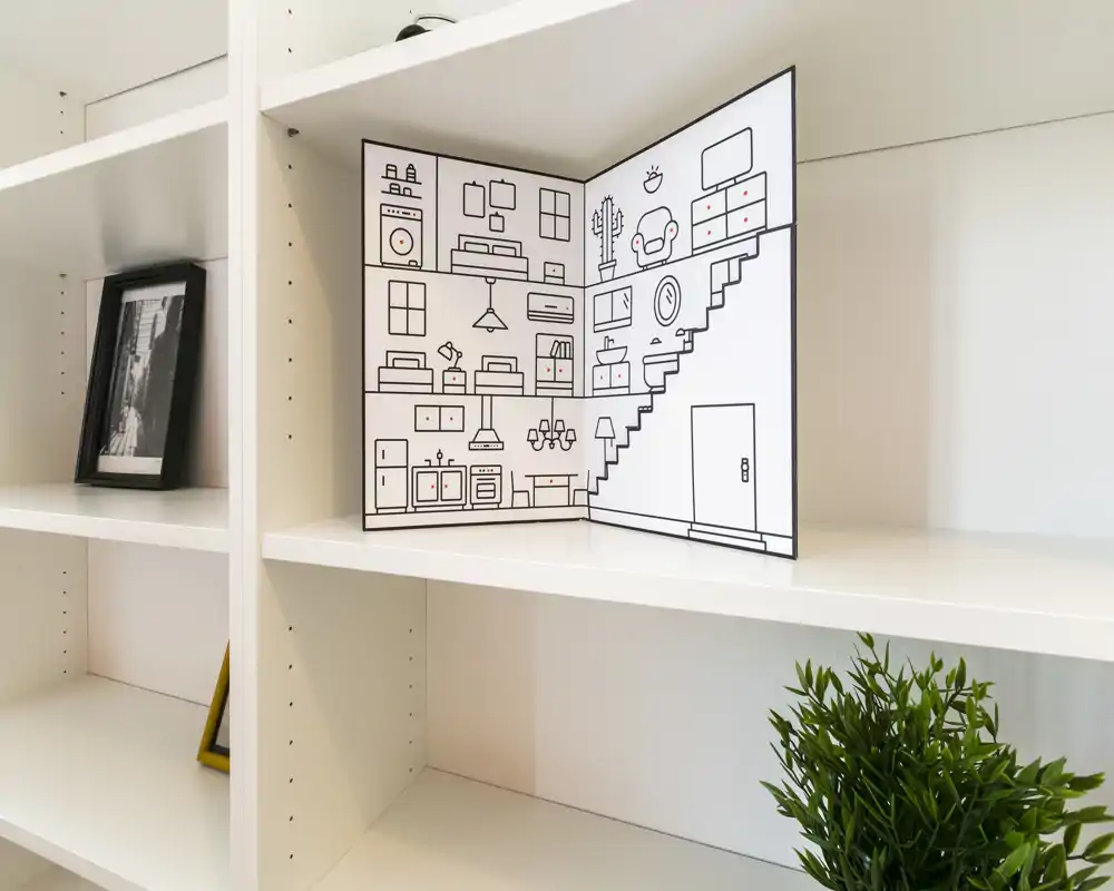 Primo piano di scaffalature bianche a giorno con un libretto decorativo raffigurante un’illustrazione di stanza, una foto incorniciata e una piccola pianta in vaso — dettaglio di scaffale, non una vista dell’intera stanza.