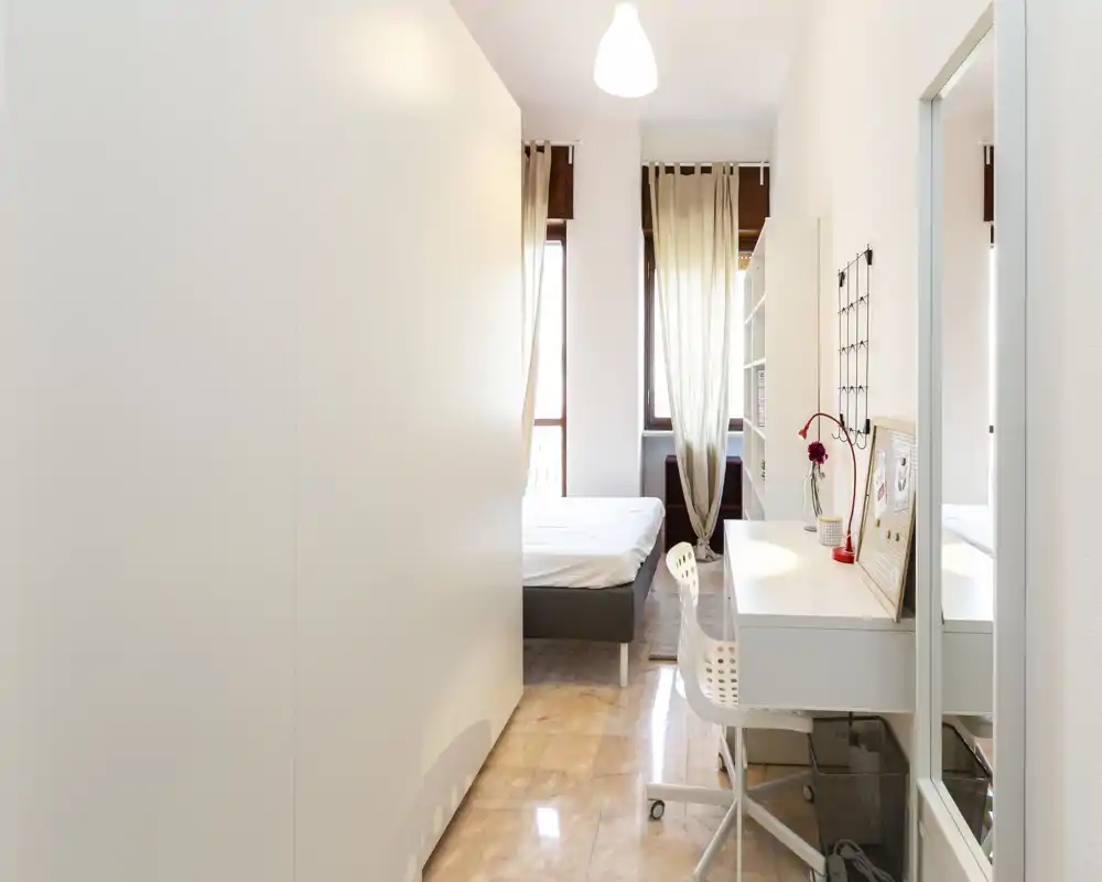 Camera da letto moderna e luminosa con letto matrimoniale visibile vicino a grandi finestre, scrivania ordinata e scaffalature; tende leggere che filtrano la luce naturale — perfetta per annunci in affitto.