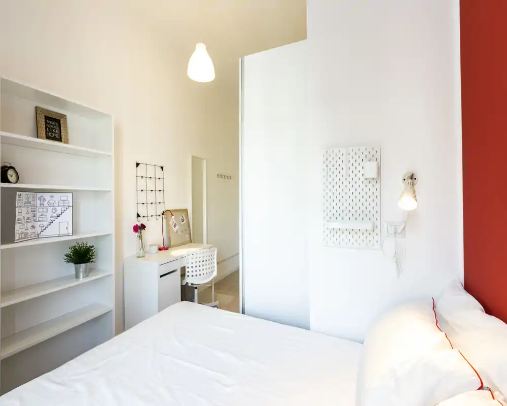 Camera da letto moderna e luminosa con letto bianco e scaffalature minimal. Una scrivania compatta e alcuni oggetti decorativi rendono l'ambiente funzionale e accogliente, con una parete rossa a contrasto.