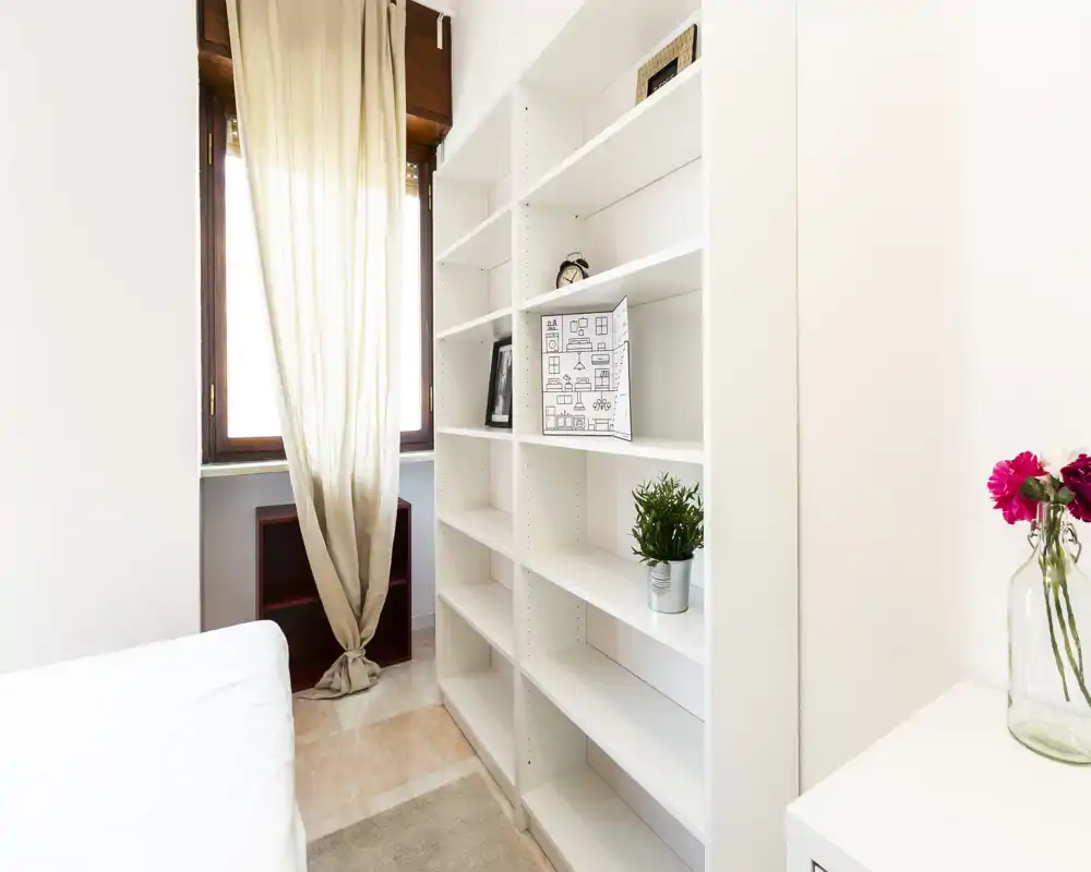 Angolo di camera luminoso e minimal con una libreria bianca, tenda leggera sulla finestra e il letto parzialmente visibile a sinistra; piante e un vaso aggiungono un leggero tocco decorativo.