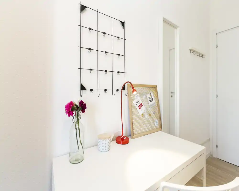 Angolo scrivania luminoso e minimal con tavolo bianco, lampada rossa, bacheca e vaso decorativo: uno spazio ordinato perfetto per studio o lavoro veloce.