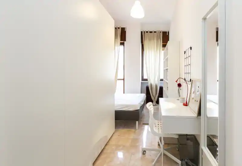 Camera da letto moderna e luminosa con letto matrimoniale visibile vicino a grandi finestre, scrivania ordinata e scaffalature; tende leggere che filtrano la luce naturale — perfetta per annunci in affitto.