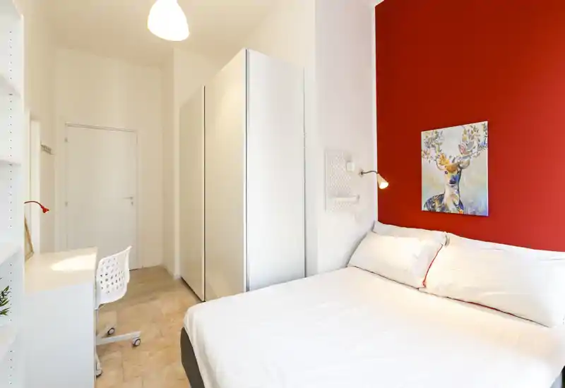 Camera da letto moderna e luminosa con parete rossa, letto matrimoniale comodo e armadio. Linee pulite, scrivania e illuminazione calda rendono la stanza accogliente per soggiorni brevi o lunghi.