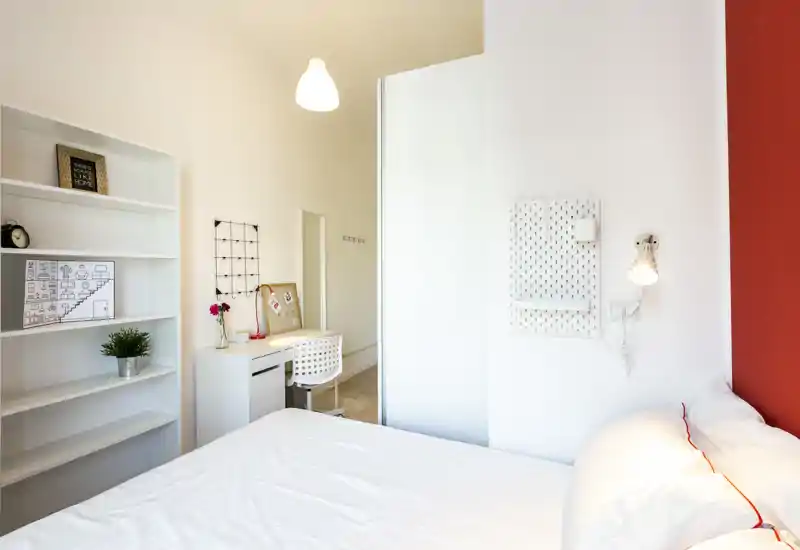 Camera da letto moderna e luminosa con letto bianco e scaffalature minimal. Una scrivania compatta e alcuni oggetti decorativi rendono l'ambiente funzionale e accogliente, con una parete rossa a contrasto.