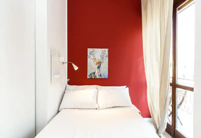 Camera da letto luminosa e di design con letto bianco su parete rossa, lampada da comodino e accesso al balcone che dona luce naturale — perfetta per annunci di affitto.