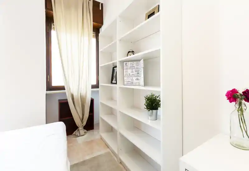 Angolo di camera luminoso e minimal con il bordo del letto visibile, grandi scaffalature bianche, una pianta in vaso e una finestra con tenda luminosa che crea un’atmosfera fresca.