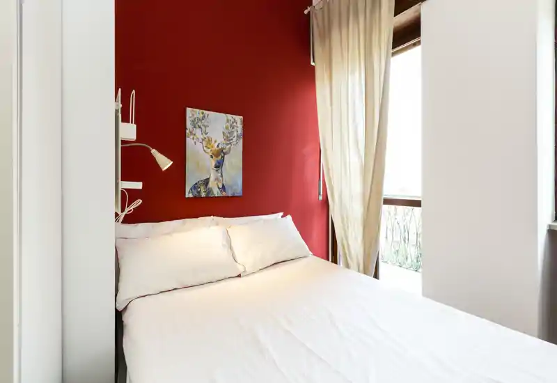 Camera da letto luminosa e ben arredata con letto matrimoniale contro una parete rossa, biancheria bianca, un quadro decorativo con cervo e luce naturale proveniente dal balcone.