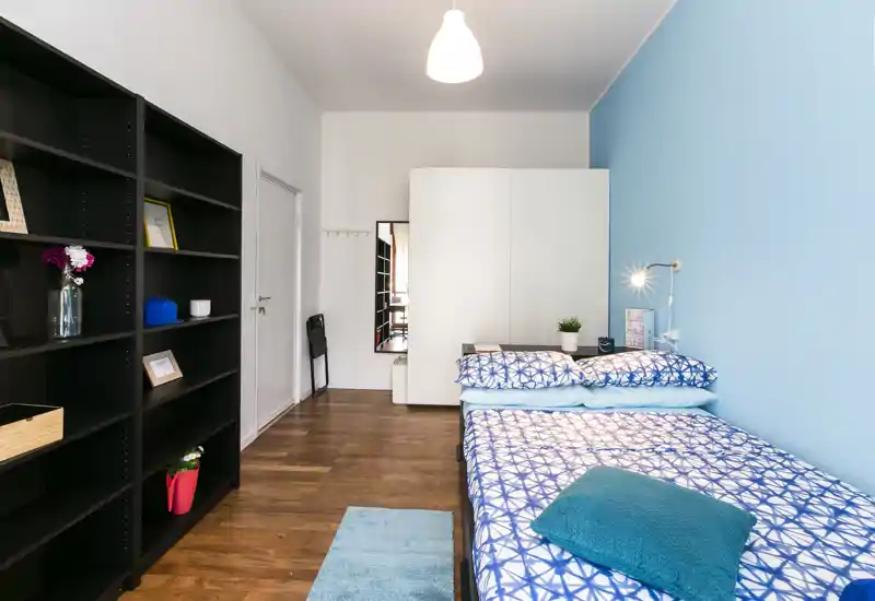 Bright, neatly styled bedroom with a double bed dressed in patterned blue linens, a bedside lamp, wardrobe and large shelving unit—ideal for rental listings highlighting storage and cozy décor.