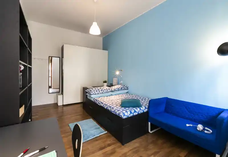 Bright, tidy bedroom with a single bed, blue accent wall and a small sofa — ideal for a comfortable stay. The space features coordinated bedding, shelving and a wardrobe, shown in a clear, well-lit photo.