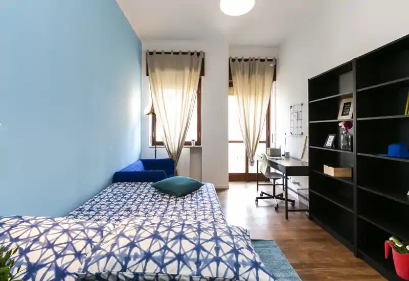 Bright, neatly staged bedroom with a patterned bed in the foreground, a small sofa and a desk by the window. Natural light and blue accent wall give the room a fresh, welcoming feel.