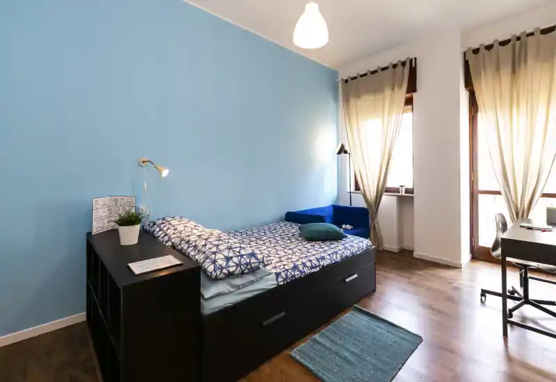 Bright, modern bedroom with a single bed against a blue accent wall, cozy textiles, a small sofa, desk by the window and plenty of natural light — ideal for a comfortable stay.