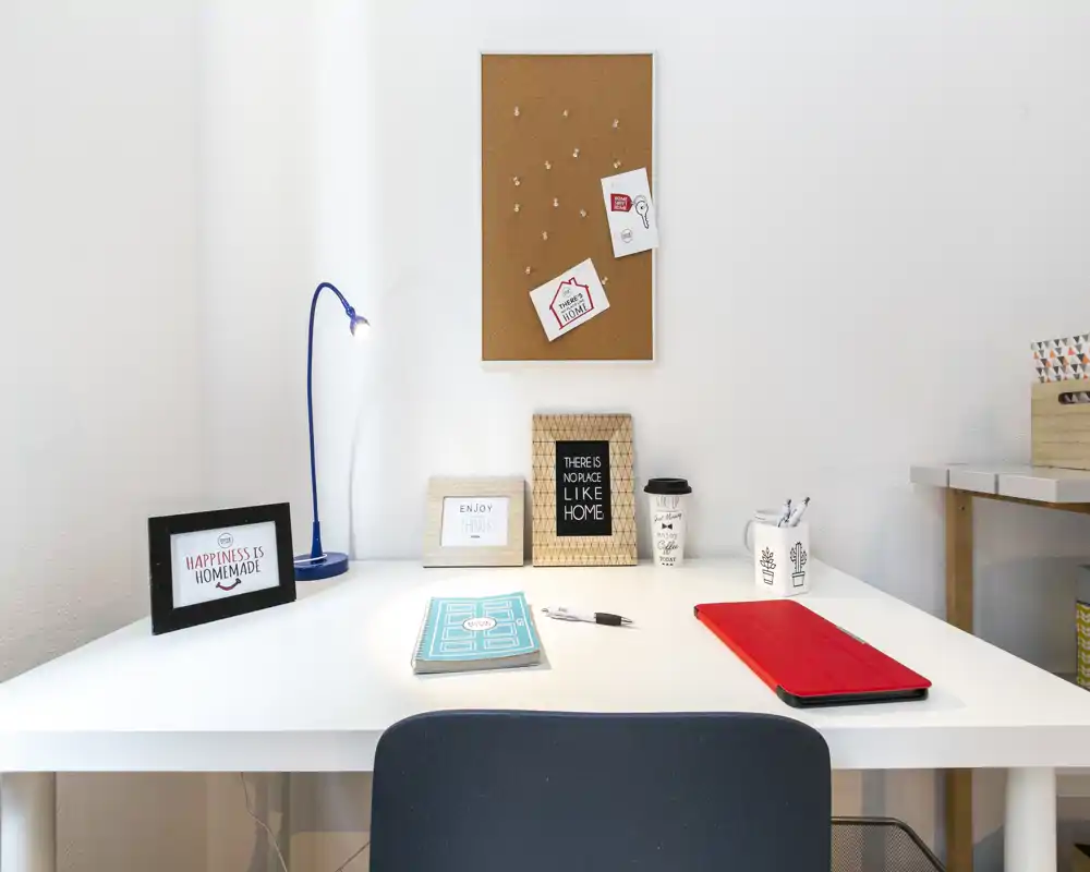 Area scrivania luminosa e ordinata con cornici, una bacheca in sughero e una lampada da lettura — un angolo lavoro accogliente in un soggiorno o studio.