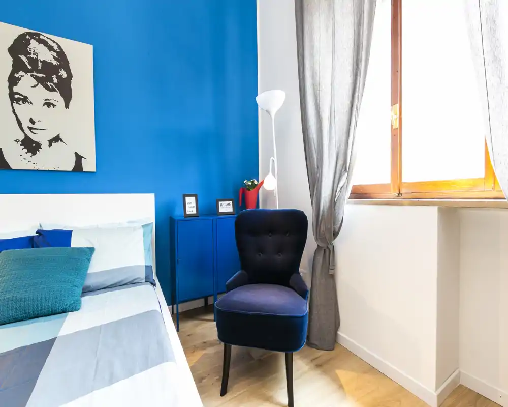 Camera da letto luminosa e alla moda con parete d'accento blu, letto comodo con cuscini sovrapposti e una poltrona accogliente vicino alla finestra.