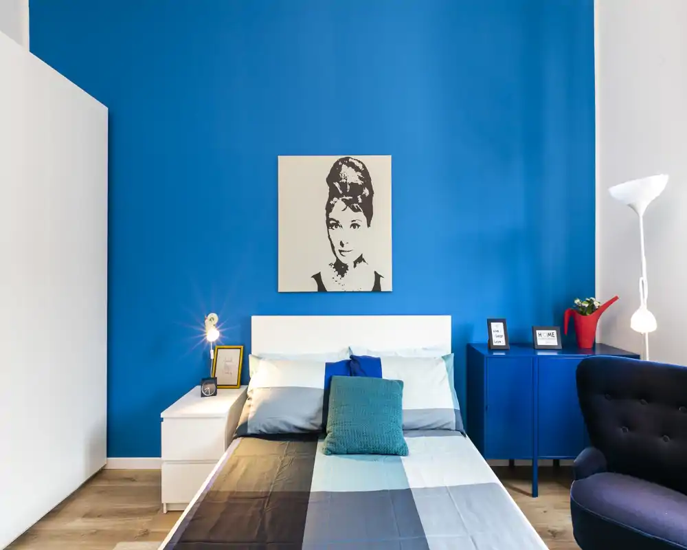 Camera da letto luminosa e di stile con parete blu, letto matrimoniale al centro con biancheria coordinata, comodini e decorazioni eleganti come quadro e lampade.