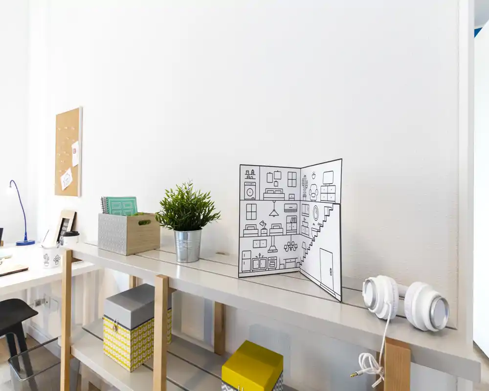 Angolo living/studio luminoso e minimal con scaffale, scatole decorative, una pianta in vaso e cuffie — area ordinata e ben illuminata ideale per uno spazio living compatto o studio.
