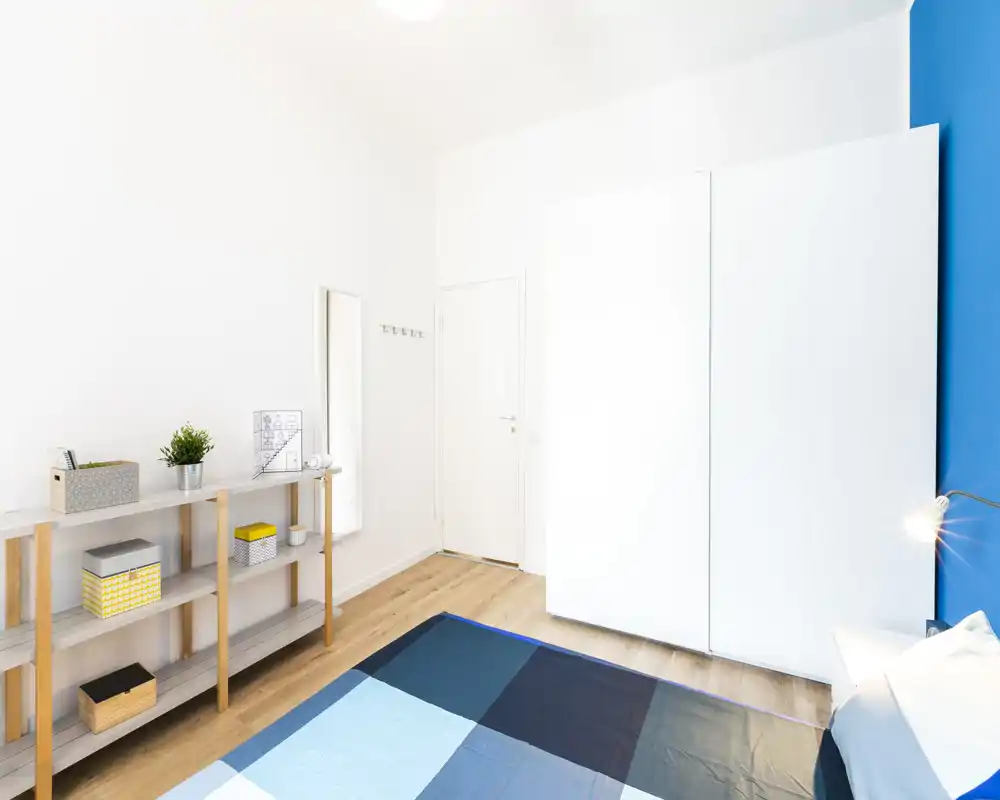 Camera da letto luminosa e moderna con letto grande e biancheria blu, armadio bianco e scaffalatura in legno — arredamento minimale e parete d’accento blu vivace.