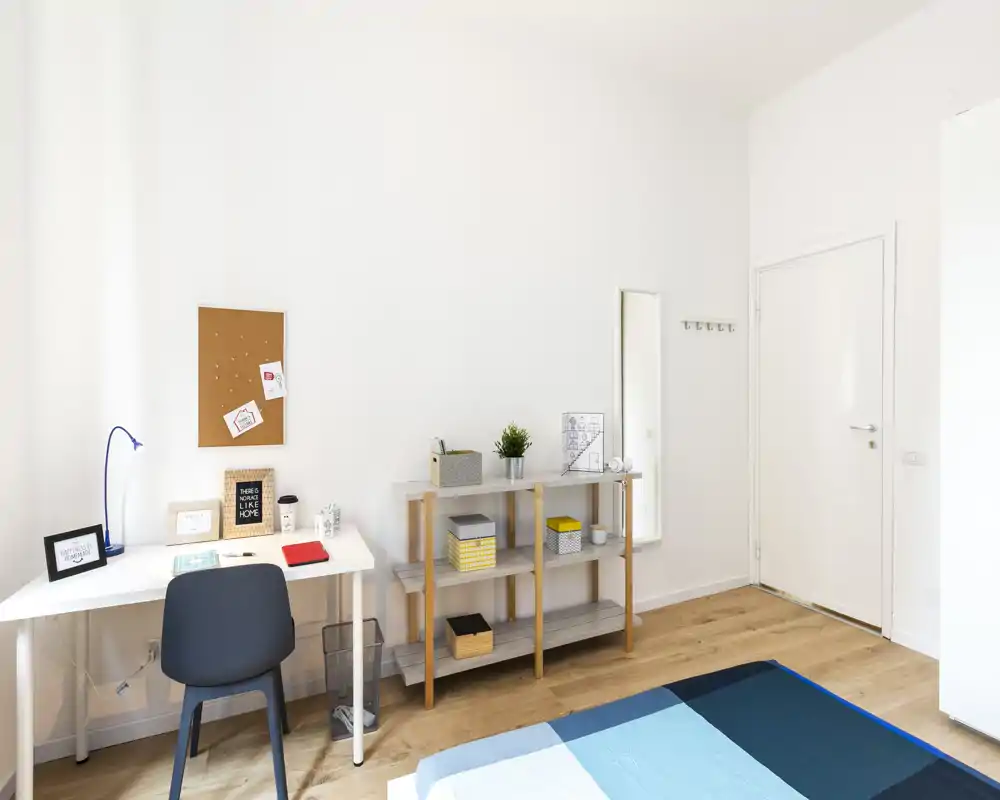 Camera da letto luminosa e minimale con bordo del letto visibile, una scrivania ordinata e scaffalature; arredo in stile nordico e luce naturale la rendono accogliente.