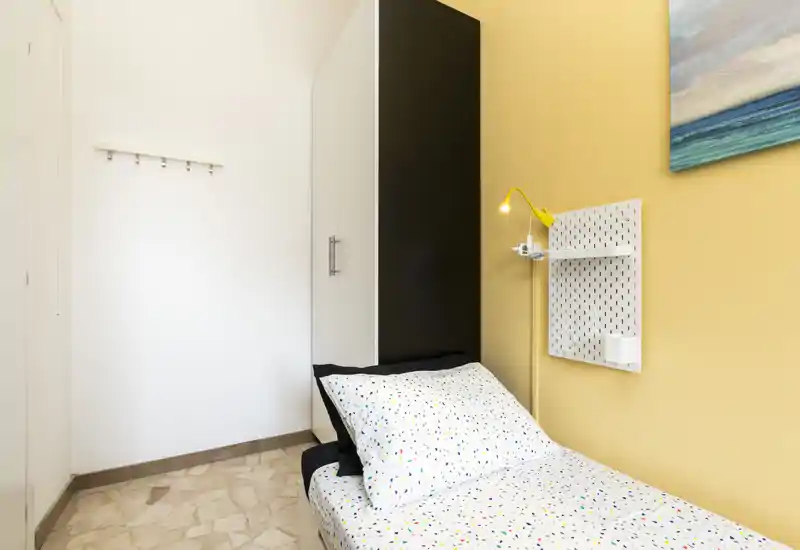 Small, neatly staged single bedroom with patterned bedding, a bedside lamp and wall storage; bright and tidy space ideal for a solo guest.