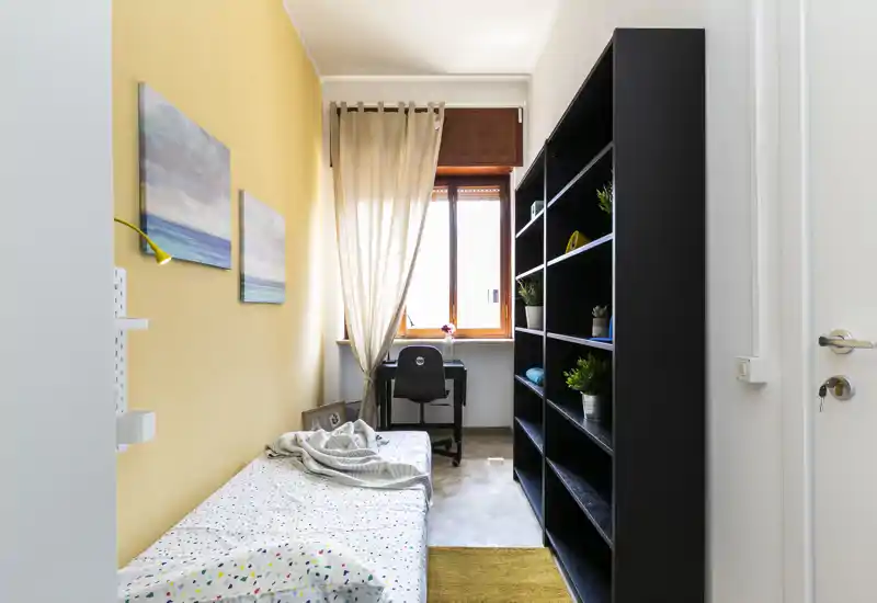 Compact bedroom with a single bed along the left wall, a desk positioned by the window and large shelving on the right. Natural light and soft textiles create a bright, comfortable space suitable for a student or single occupant.