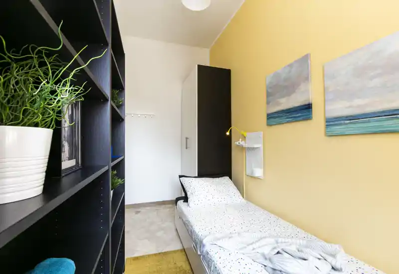 Compact single bedroom with a neatly made bed, tall wardrobe and open shelving; warm yellow wall and coastal artwork create a welcoming atmosphere.