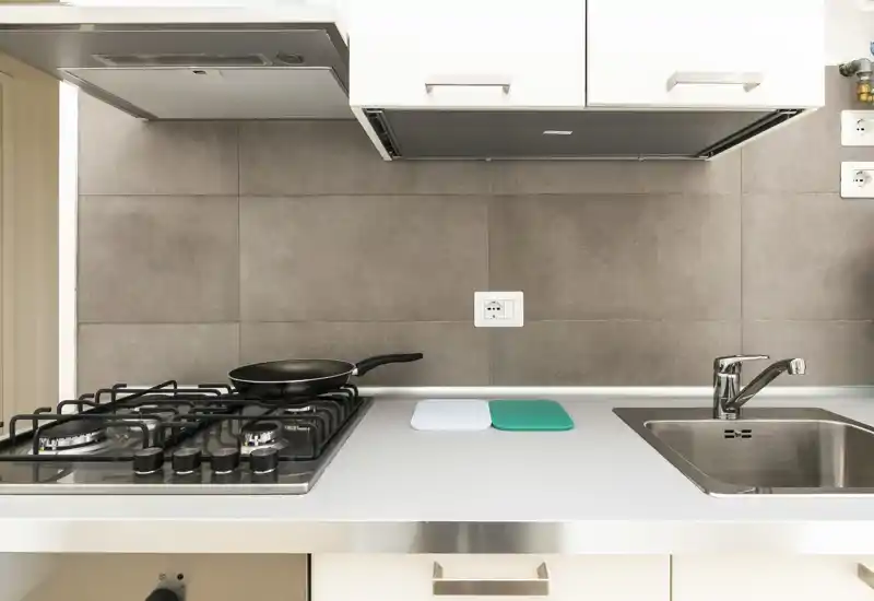 Angolo cucina moderno e compatto con piano cottura a gas, lavello in acciaio e schienale piastrellato neutro. Piano di lavoro pulito con pochi accessori (padella, taglieri) e illuminazione uniforme e luminosa.
