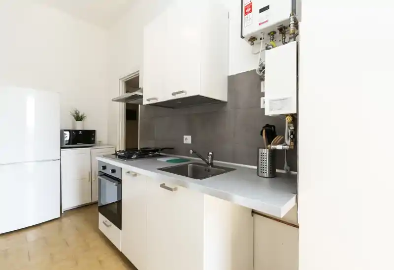 Cucina compatta moderna e luminosa con mobili bianchi, piano cottura a gas, forno incassato e frigorifero; piano lavoro pulito con lavello e porta-utensili.