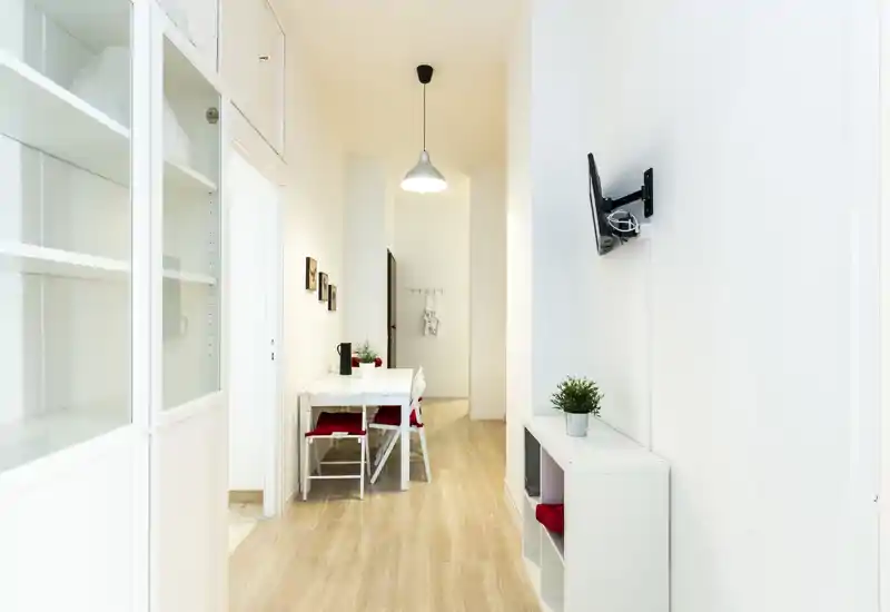 Zona giorno luminosa e minimal con un piccolo tavolo da pranzo e scaffalature semplici; pareti bianche, pavimento chiaro e piante decorative creano un ambiente arioso e accogliente.