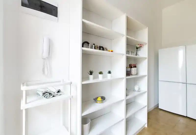 Angolo cucina luminoso e compatto con scaffali bianchi aperti, piccole piante decorative e barattoli, e un frigorifero bianco — stile moderno essenziale, adatto per annunci.