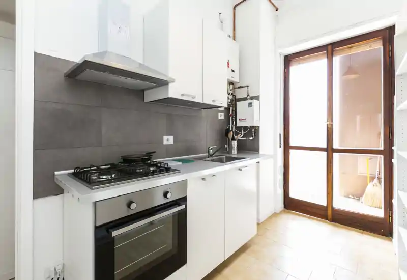 Cucina compatta con piano cottura a gas, forno integrato e lavello; luminosità naturale grazie alla porta finestra che apre su un piccolo balcone.