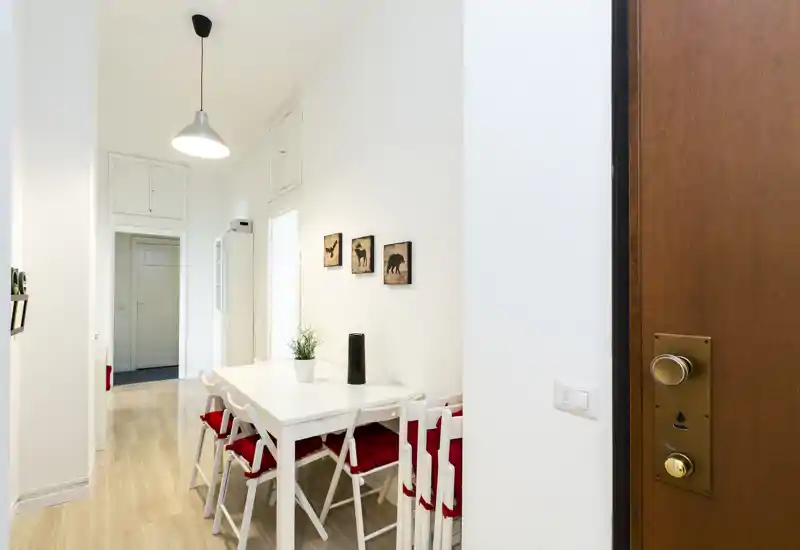 Zona pranzo luminosa e compatta con tavolo bianco e sedie pieghevoli con cuscini rossi, quadretti minimal sulle pareti e stile pulito e nordico — ideale per pasti raccolti.