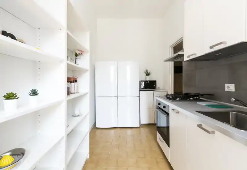 Cucina moderna e luminosa con mobili bianchi, frigorifero a doppia anta, forno incassato, piano cottura a gas e scaffalatura aperta—pulita, minimal e ben organizzata.