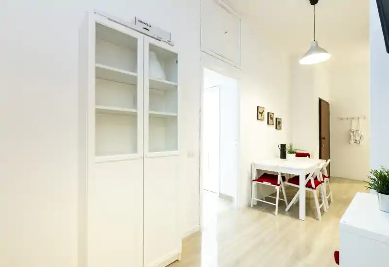 Zona pranzo compatta e luminosa con tavolo bianco semplice, quattro sedie con cuscini rossi e pochi elementi decorativi, perfetta per pasti accoglienti in un appartamento moderno.
