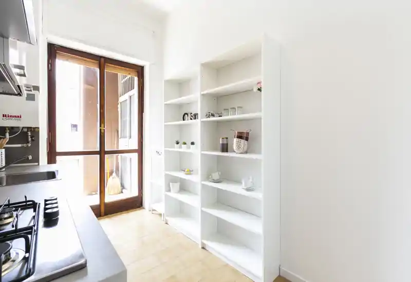 Cucina compatta e luminosa con piano cottura a gas, lavello in acciaio e scaffalature a vista; luce naturale da una porta finestra che illumina il pavimento piastrellato e lo spazio essenziale.