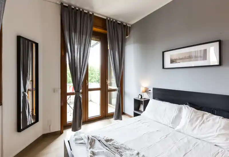 Camera da letto moderna e luminosa con un grande letto matrimoniale, comodino e porta finestra verso un piccolo balcone; tonalità neutre e tessuti morbidi creano un'atmosfera rilassante.