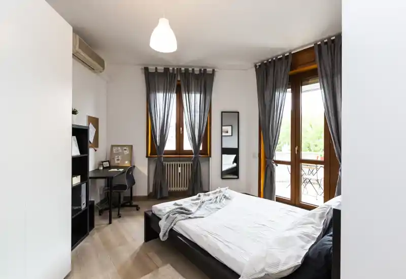 Camera da letto luminosa e ben arredata con un ampio letto matrimoniale, angolo studio e porta sul balcone che garantisce molta luce naturale—ideale per chi cerca una stanza moderna e confortevole.