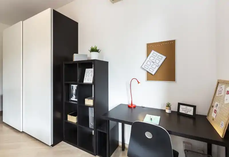 Angolo camera ordinato adibito a piccolo spazio di lavoro con scrivania nera, scaffale e armadio. Stile minimale e luminoso con una lampada rossa, bacheca e piante per un aspetto moderno e funzionale.