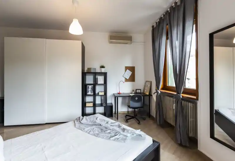 Camera da letto luminosa e moderna con letto matrimoniale, armadio, scrivania e ampia finestra con tende; ambiente ben illuminato e accogliente.