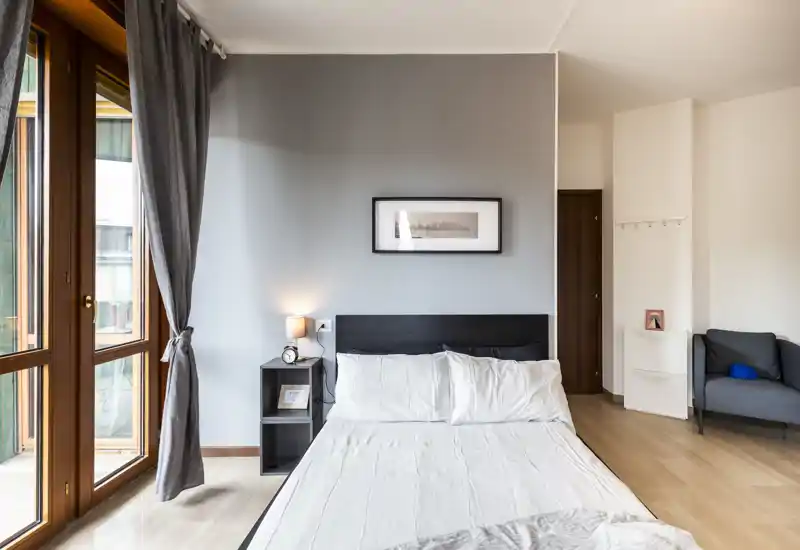 Camera da letto moderna e luminosa con letto matrimoniale, comodino e grandi finestre a tutta altezza che lasciano entrare molta luce naturale.