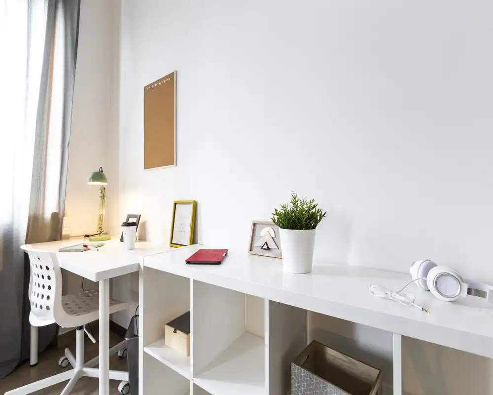 Angolo studio luminoso e minimale con una lunga scrivania bianca, scaffalatura, sedia da scrivania e dettagli decorativi come una pianta in vaso e foto incorniciate — perfetto per lavorare concentrati.