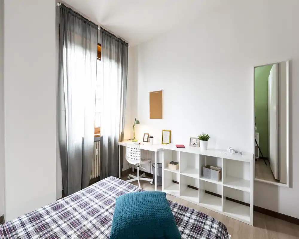 Camera da letto moderna e luminosa con copriletto a quadri, cuscino turchese e scrivania vicino alla finestra. La luce naturale riempie l'ambiente e gli scaffali bianchi con decorazioni minimal danno una sensazione ariosa.
