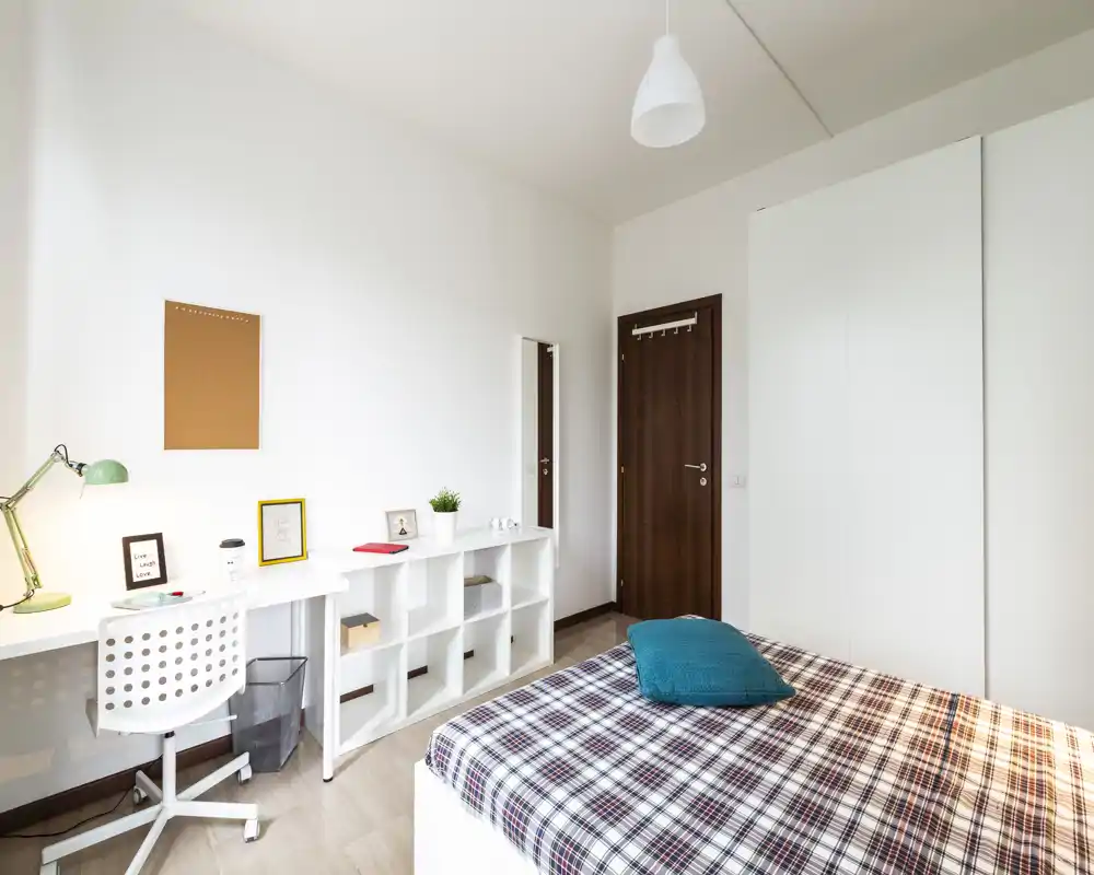 Camera da letto luminosa e moderna con letto matrimoniale coperto da una coperta a quadri, un cuscino turchese e una scrivania con scaffalatura a parete. Pareti bianche pulite, armadio a muro e luce naturale rendono lo spazio arioso e funzionale, ideale per studenti o professionisti.