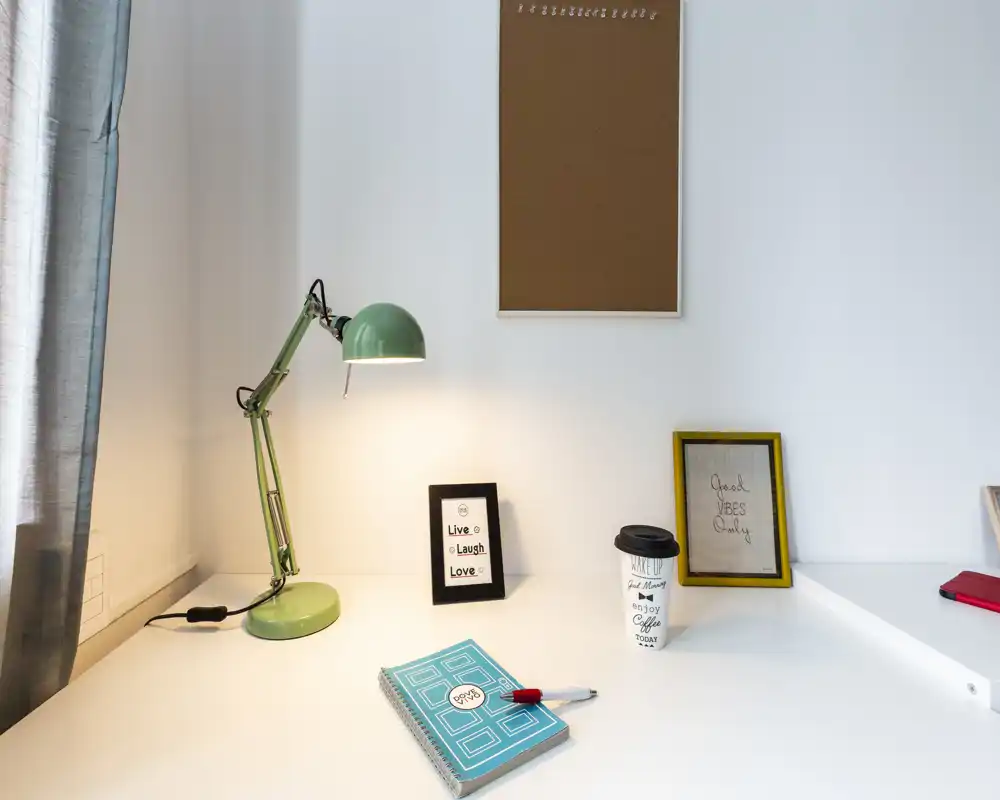 Angolo scrivania luminoso e ordinato con una lampada verde, cornici e un quaderno — ideale per lavorare o studiare in un soggiorno compatto.