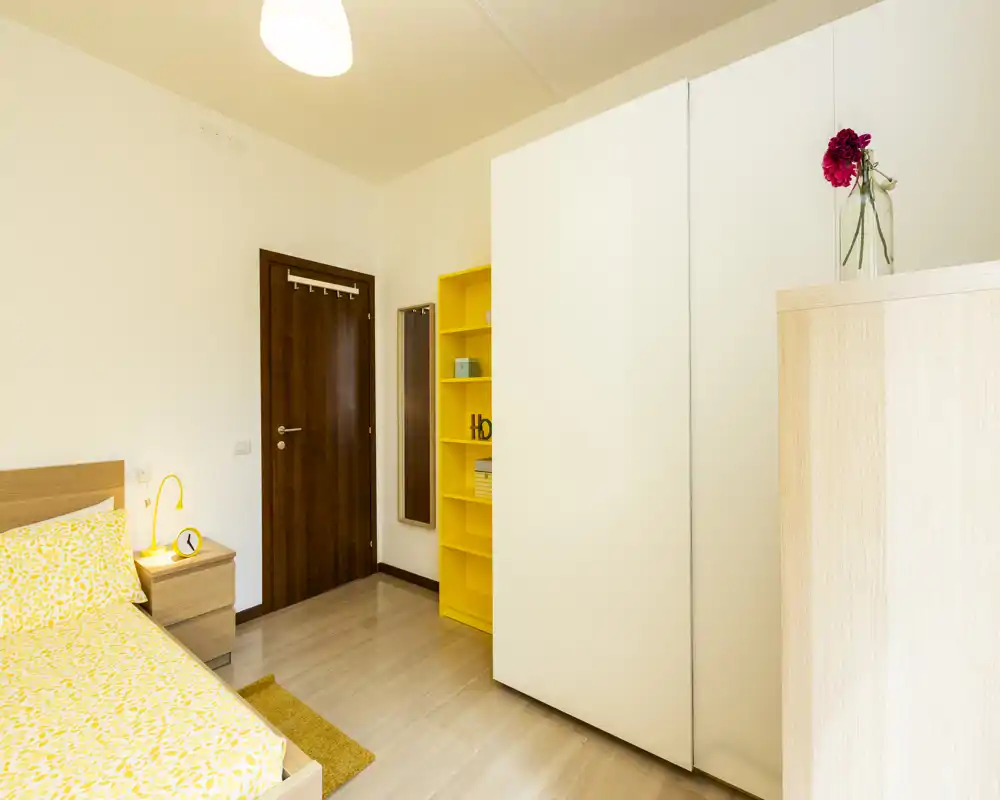 Chambre moderne et lumineuse avec un lit simple bien fait, table de chevet et armoire. Palette neutre avec des touches jaunes pour une atmosphère fraîche et accueillante, idéale pour une annonce locative.