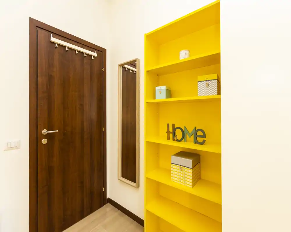 Entrée lumineuse et moderne avec porte en bois, miroir pleine hauteur et une étagère jaune vive pour rangement et décoration.